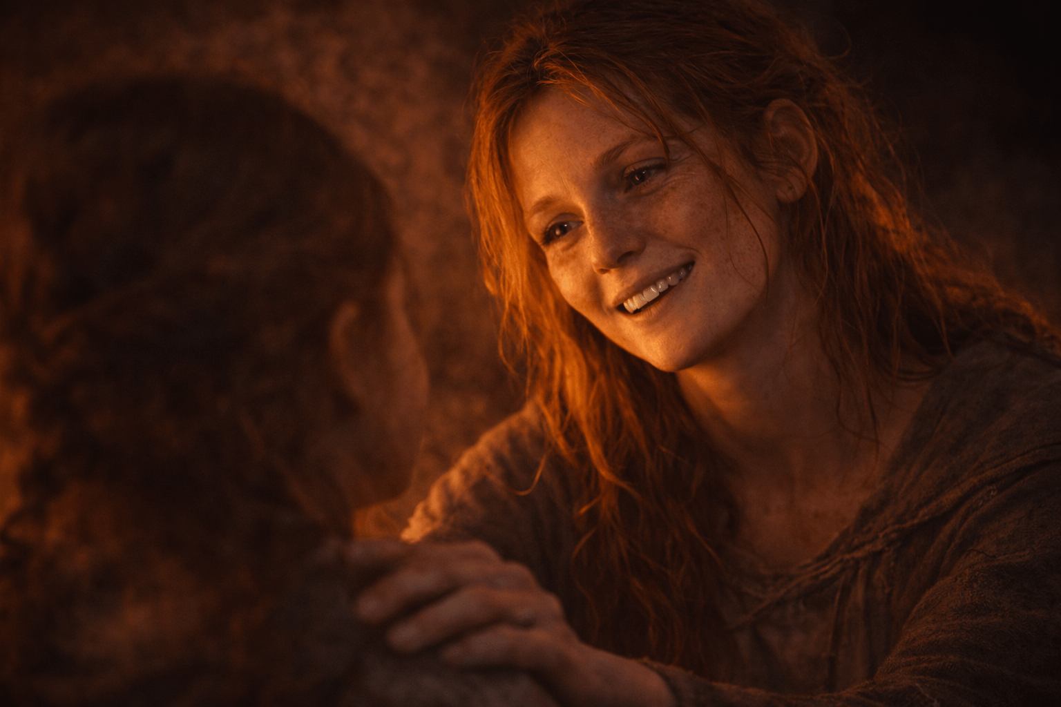 Red-haired woman with freckles smiles tenderly, hand on another person's shoulder, conveying comfort and reassurance.