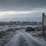 A weathered welcome sign for Iqaluit stands on a snowy, desolate road leading toward a distant, frozen coastal town.