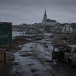 A rusted welcome sign for Prince Edward Island stands beside a desolate, debris-strewn road leading to a ruined coastal village under a grey sky.