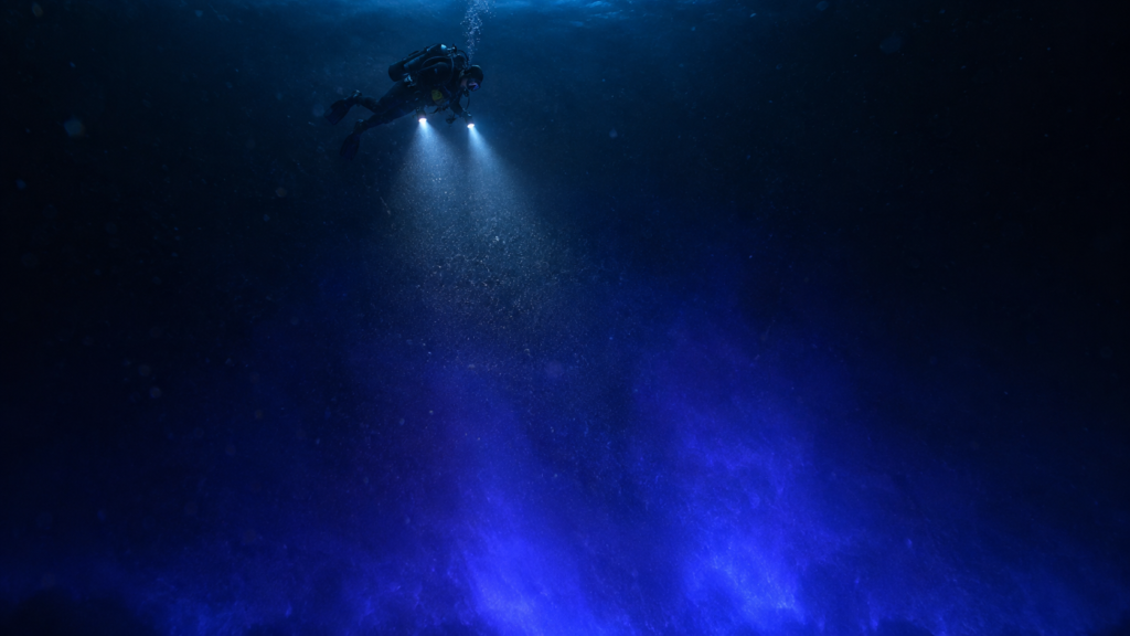 A scuba diver swims through dark, deep blue water, illuminating the surrounding area with two bright underwater lights.