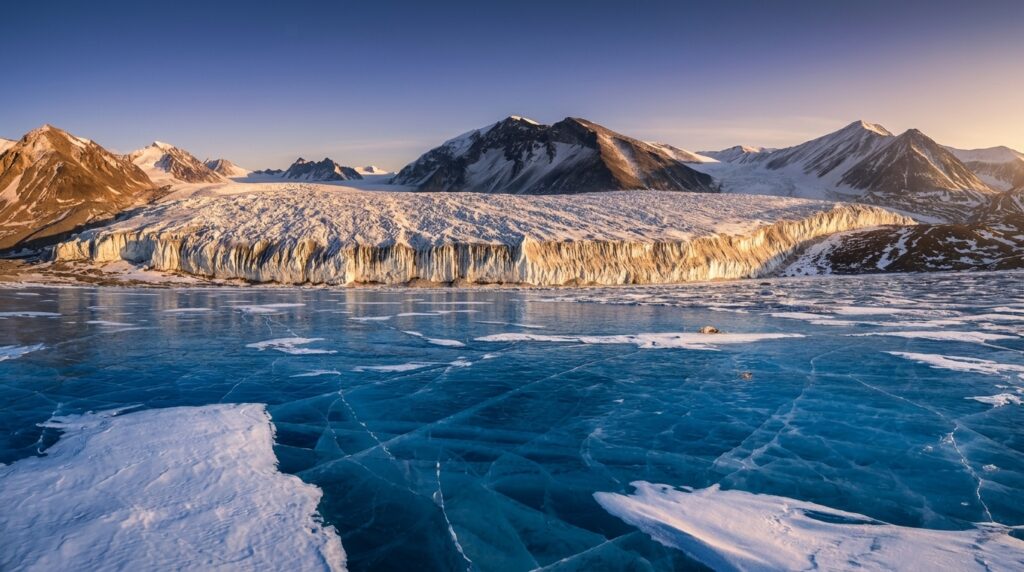 A vast, rugged glacier sits before a range of snow-capped mountains under a clear, pale sky, with a foreground of cracked, deep blue ice.