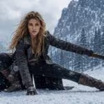 A woman in weathered post-apocalyptic gear crouches in a snowy mountain valley, holding a large axe.